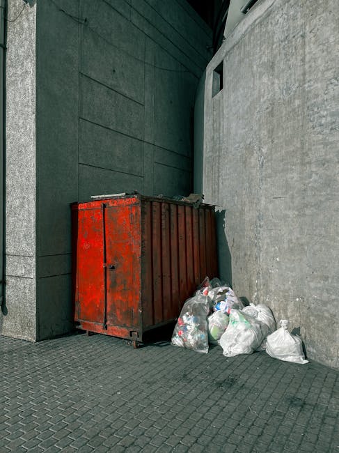A pile of discarded cardboard boxes, some flattened and others partially intact, is situated against a weathered brick and concrete wall. The boxes vary in size and are primarily brown with printed la
