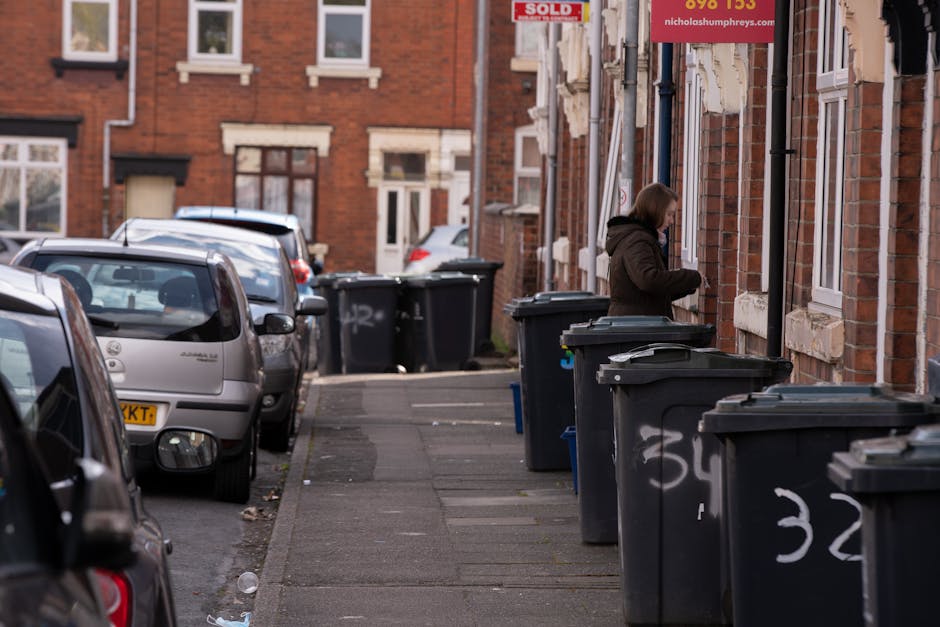 A woman wearing a dark brown jacket with a hood stands on a pavement alongside a row of large black wheelie bins with closed lids, positioned against the brick exterior wall of a residential building.