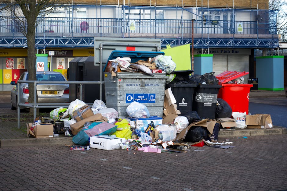 A large cluster of mixed waste and rubbish bags overflowing from multiple bins and containers, situated on a paved pavement area near a parking spot. The refuse includes cardboard boxes, paper, plasti