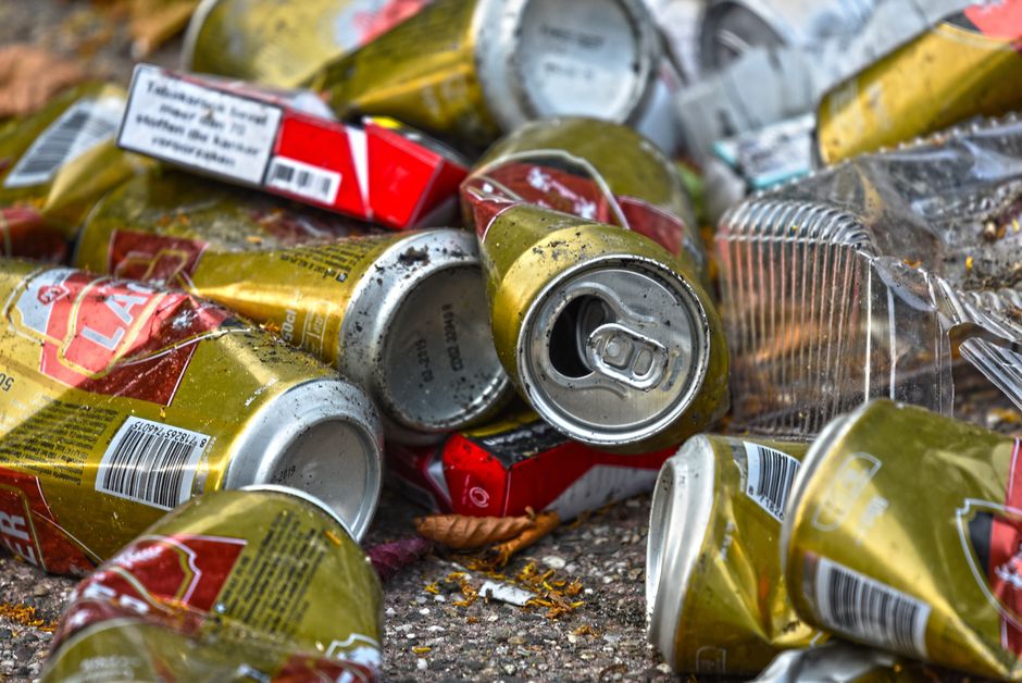 A large cluster of mixed waste and rubbish bags overflowing from multiple bins and containers, situated on a paved pavement area near a parking spot. The refuse includes cardboard boxes, paper, plasti